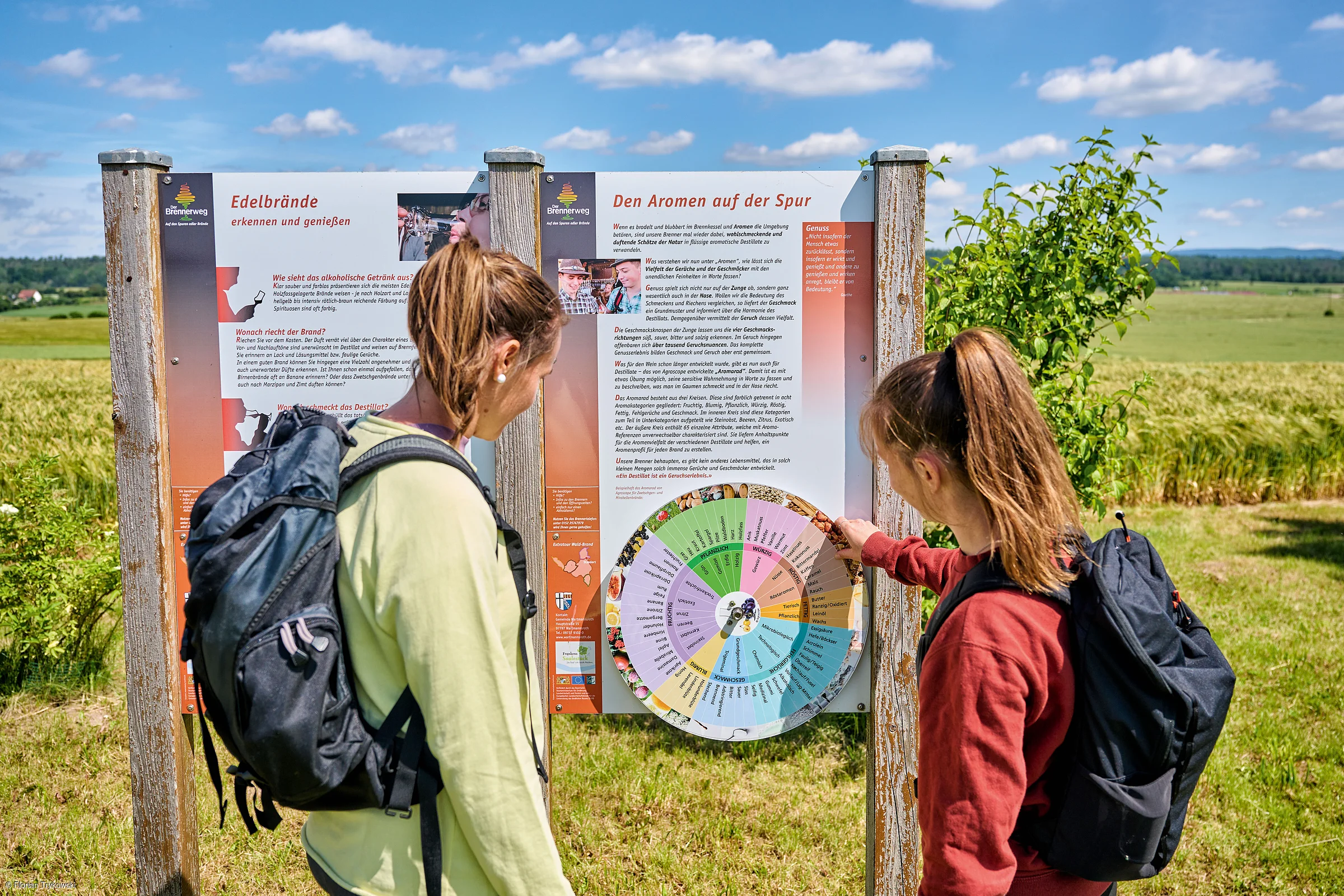Zwei Frauen mit Rucksäcken lesen eine Infotafel über Edelbrände in einer ländlichen Umgebung bei Sonnenschein.