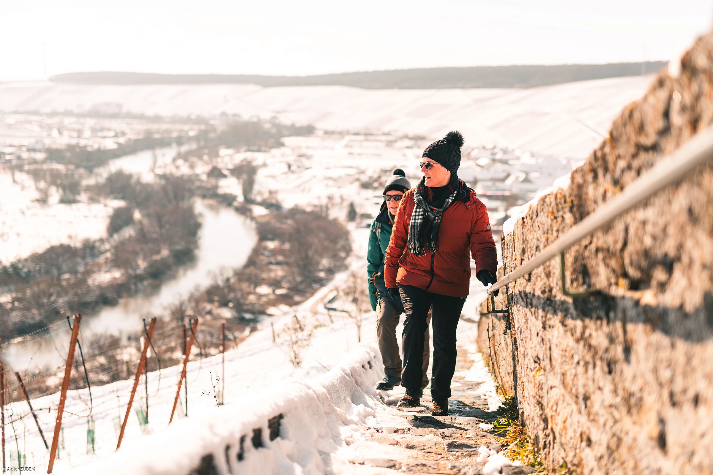 Zwei Personen in Winterkleidung gehen einen schneebedeckten Weg neben einer Steinmauer bergauf.