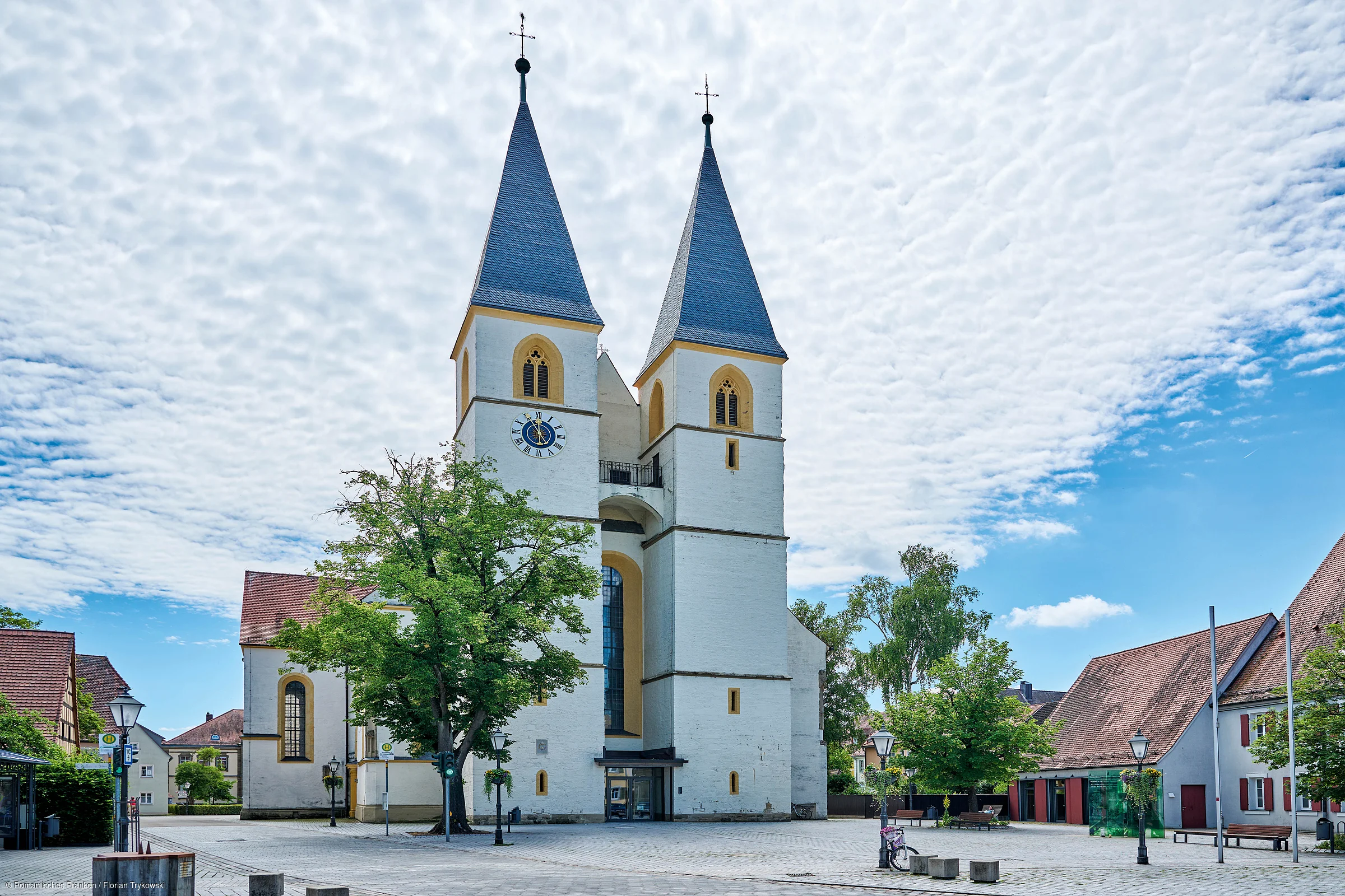 Kirche mit zwei hohen Türmen, Uhr, Bäumen und umliegenden Gebäuden unter bewölktem Himmel.