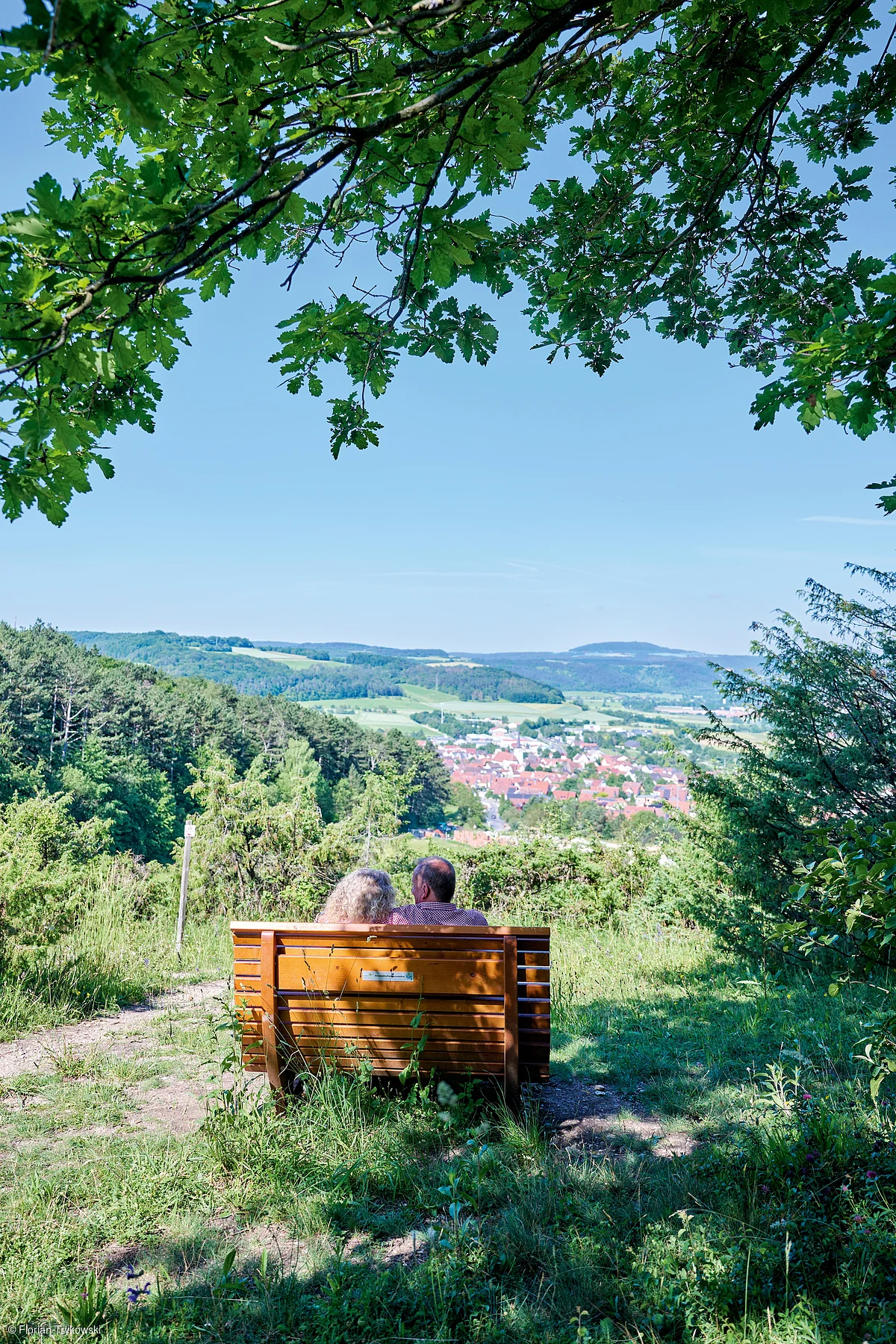 Paar sitzt auf Holzbänken mit Blick auf eine Stadt, umgeben von Bäumen und Wiesen unter blauem Himmel.