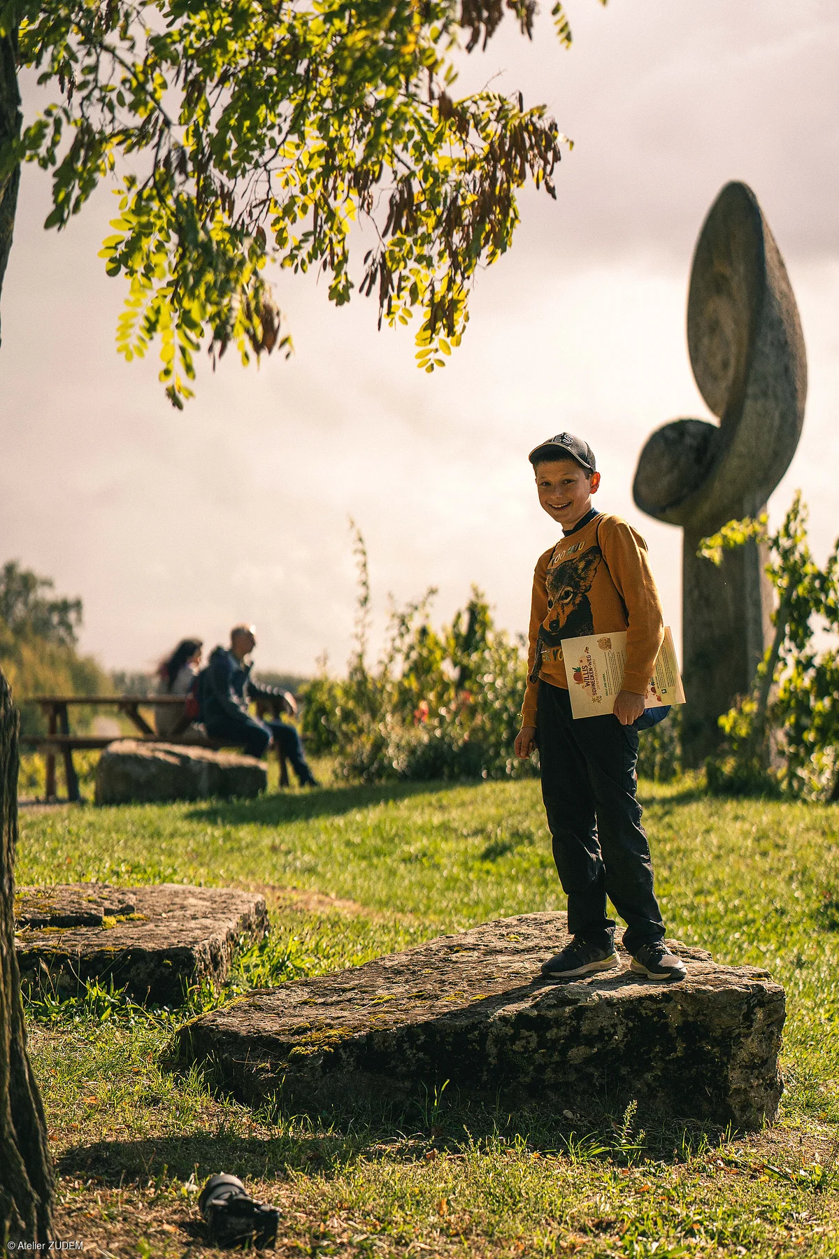 Junge mit Mütze steht auf Steinplatte im Park, hält Papier, im Hintergrund zwei sitzende Personen.