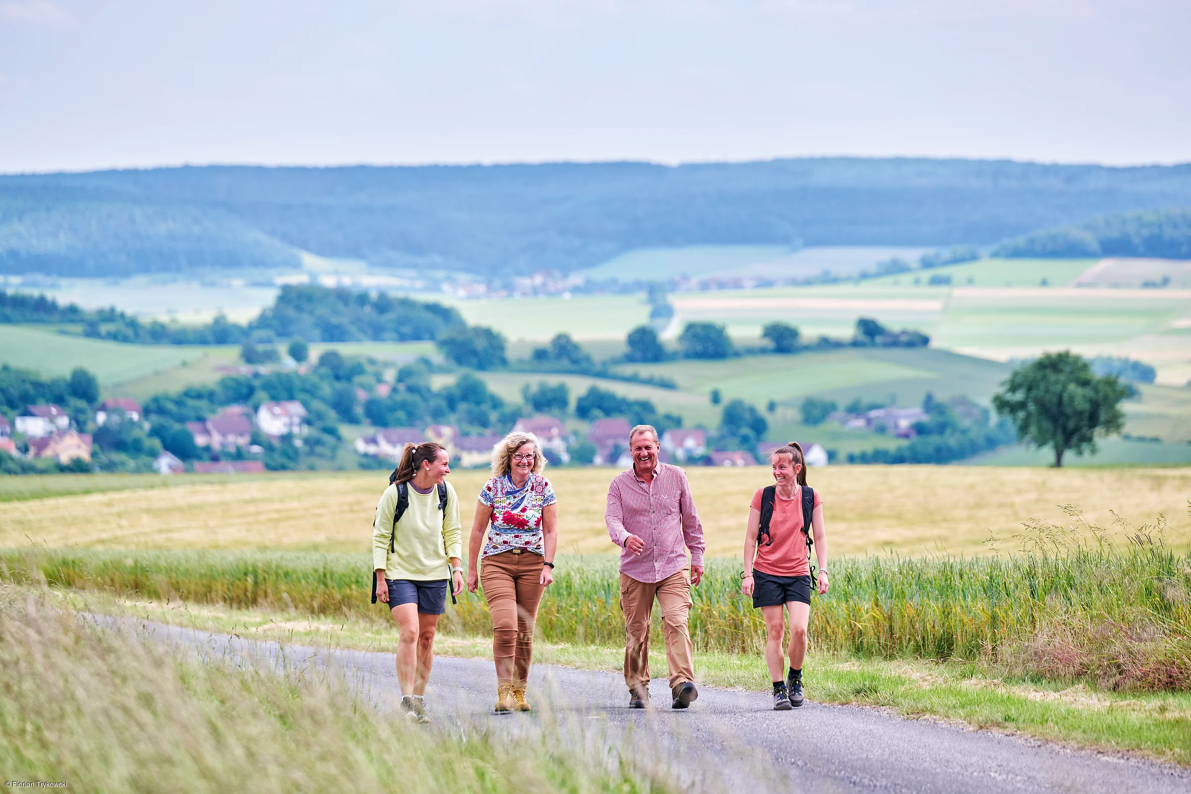 Vier Personen wandern auf einem Feldweg mit grünen Feldern und Hügeln im Hintergrund.