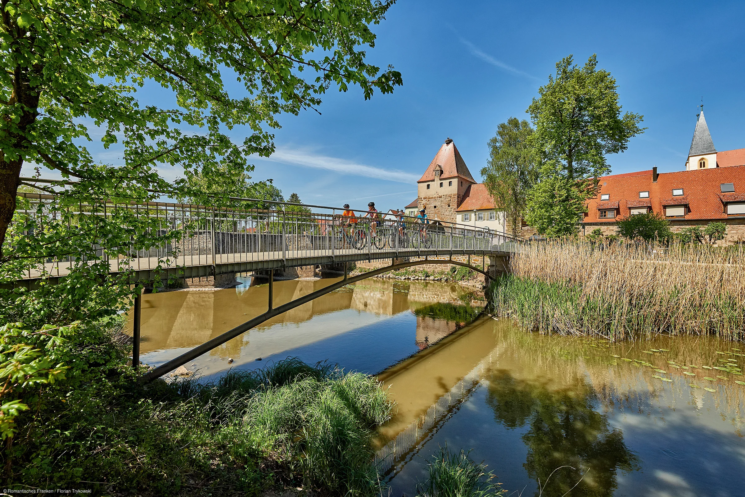 Brücke über Fluss mit Radfahrern, Bäumen und historischen Gebäuden bei klarem Himmel im Hintergrund.