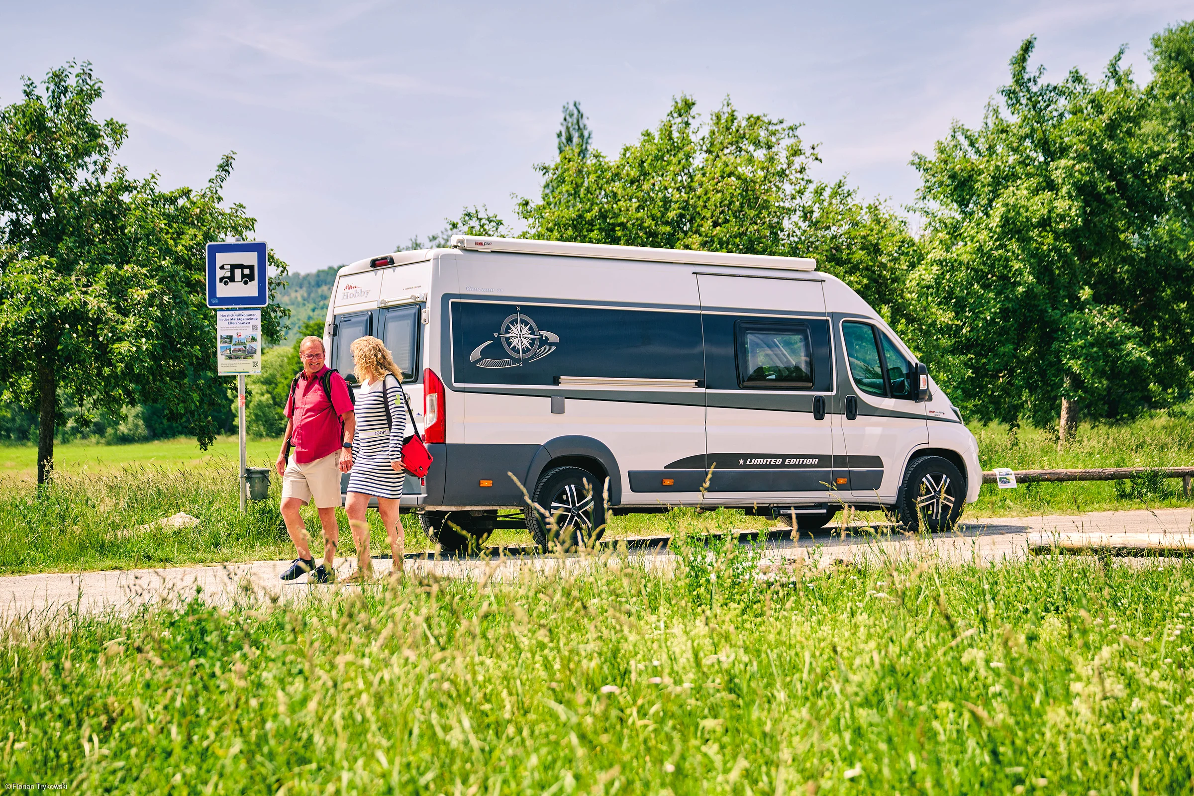 Zwei Personen gehen an einem geparkten Wohnmobil auf einem Weg entlang, umgeben von grüner Wiese und Bäumen.