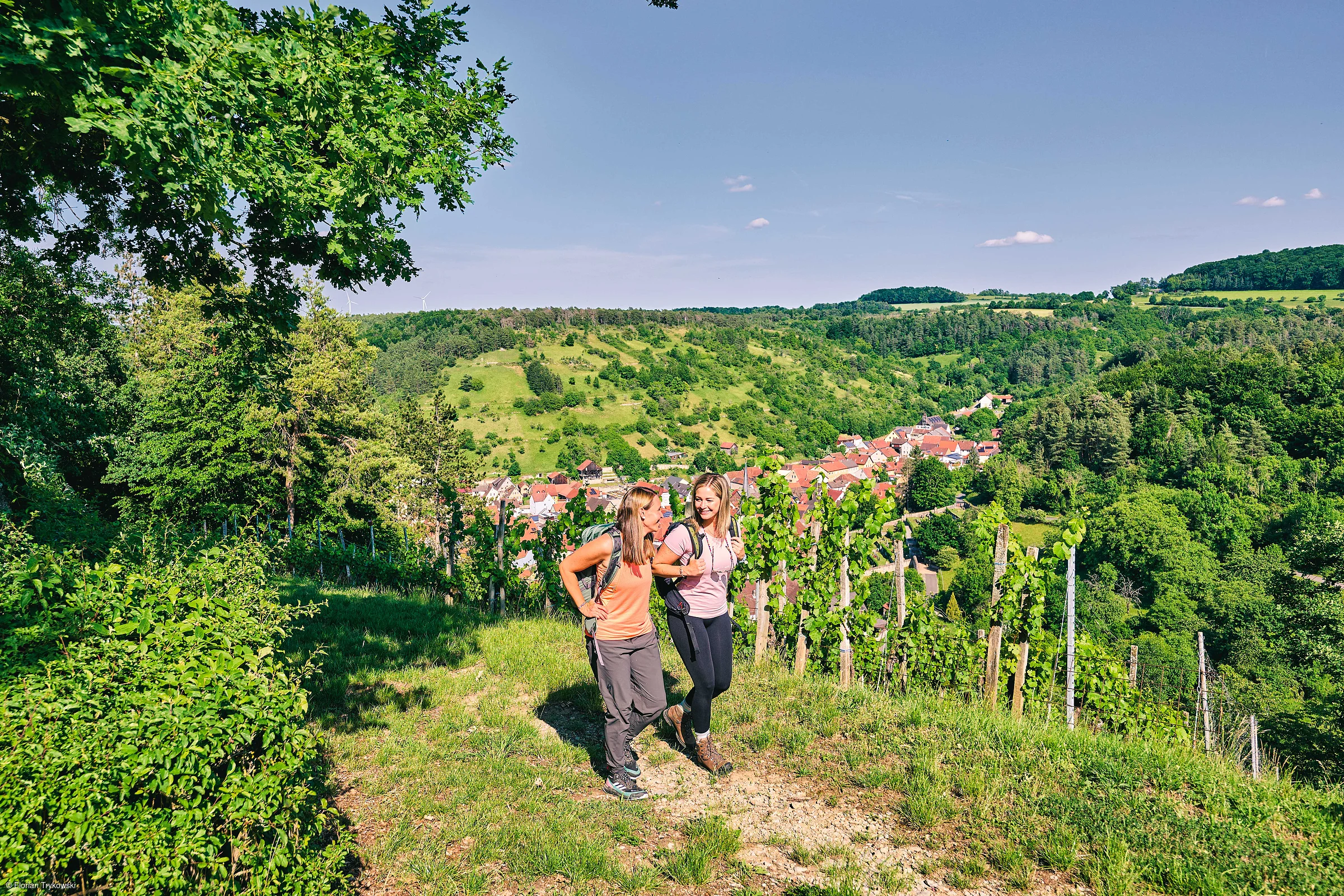 Zwei Frauen mit Rucksäcken wandern auf einem Weg mit Blick auf ein Dorf und bewaldete Hügel im Hintergrund.