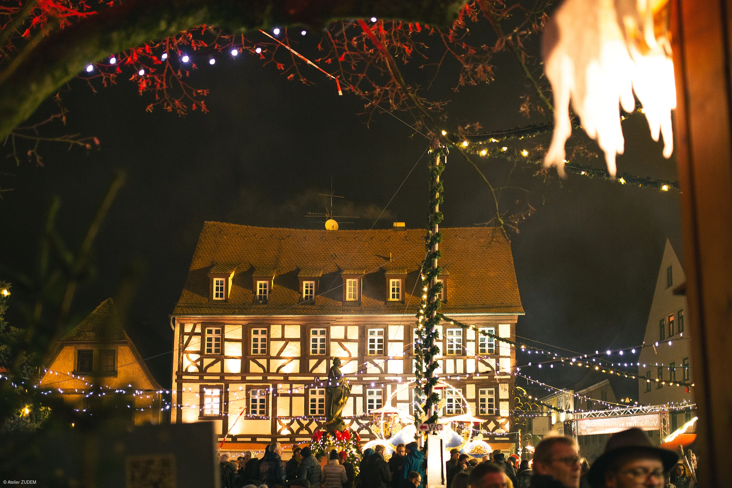 Beleuchtetes Fachwerkhaus bei Nacht mit Menschen und Weihnachtsbeleuchtung auf einem Markt.
