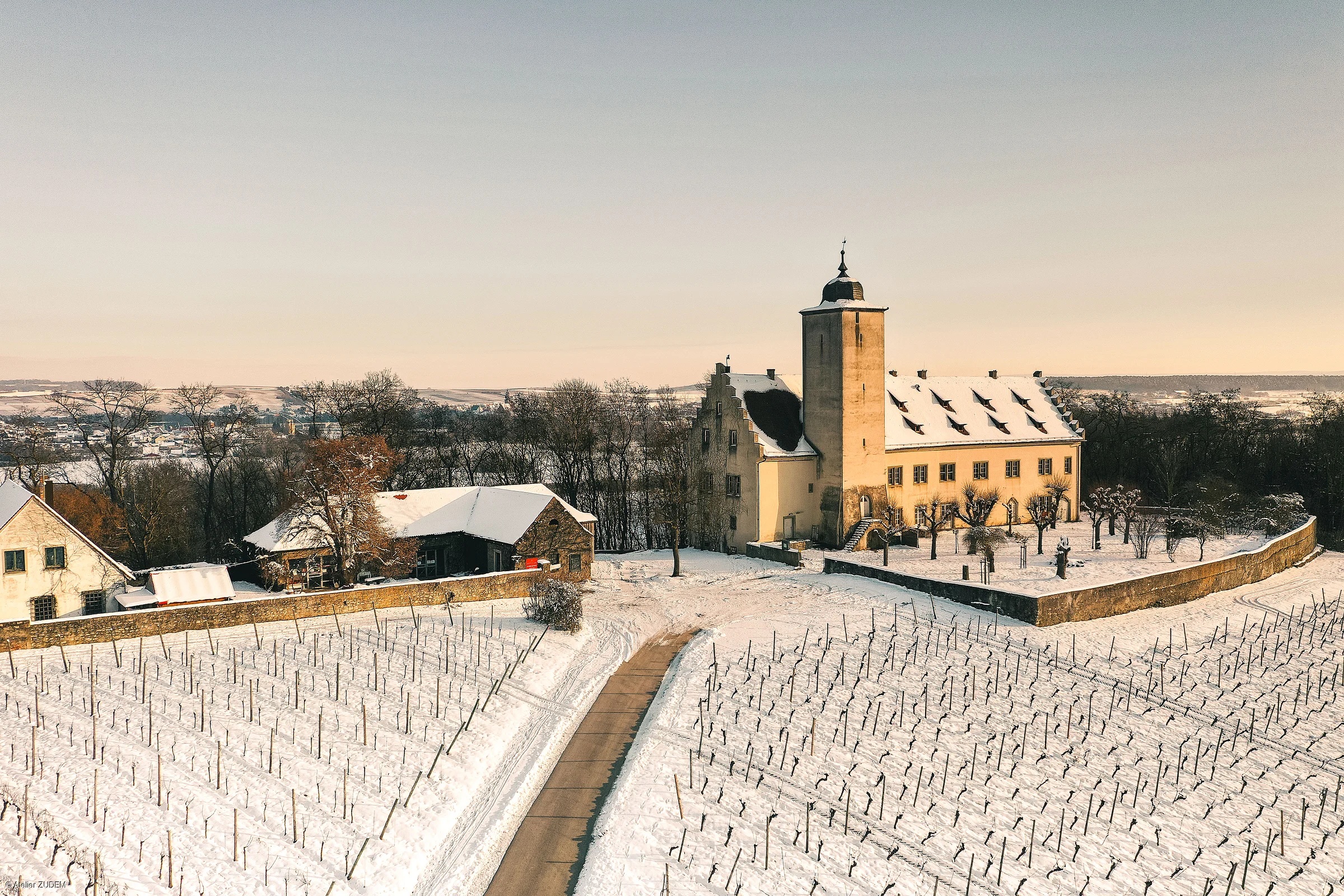 Schneebedecktes Schloss mit Turm und umliegenden Weinbergen im Winter bei Sonnenuntergang.