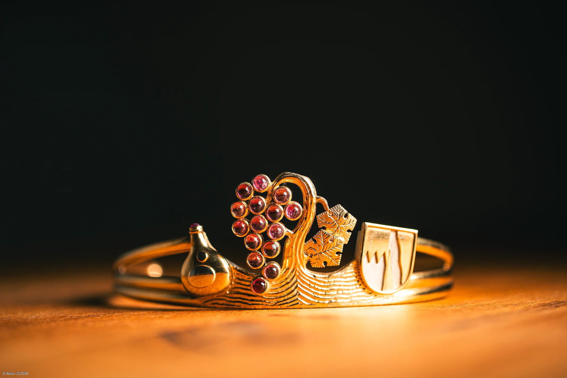 Goldener Armreif mit Trauben, Weinflasche, Weinblättern und Wappen auf Holztisch vor schwarzem Hintergrund