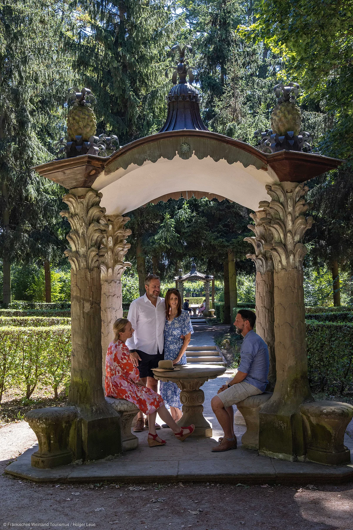 Vier Personen sitzen und stehen an einem runden Steintisch unter einem verzierten Pavillon im Park.