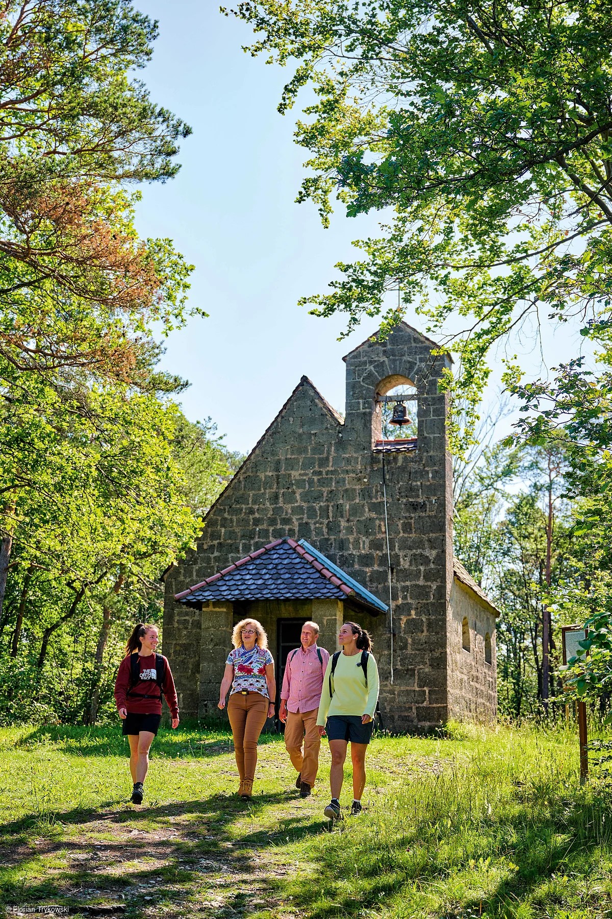 Vier Wanderer gehen auf einem Waldweg vor einer kleinen Steinkapelle mit Glockenturm bei Sonnenschein.