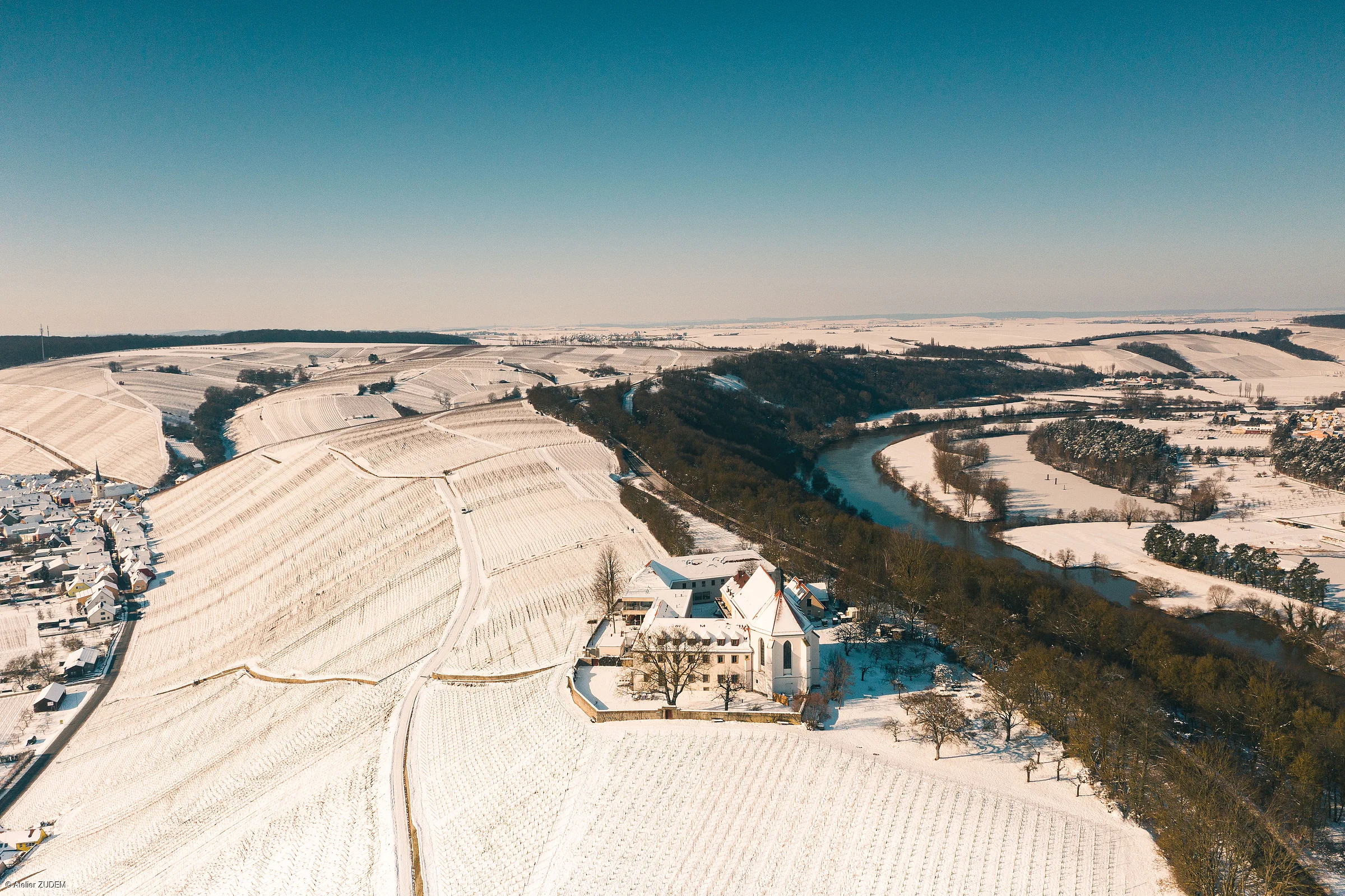 Luftaufnahme einer verschneiten Landschaft mit Fluss, Feldern und Gebäuden unter klarem Himmel.