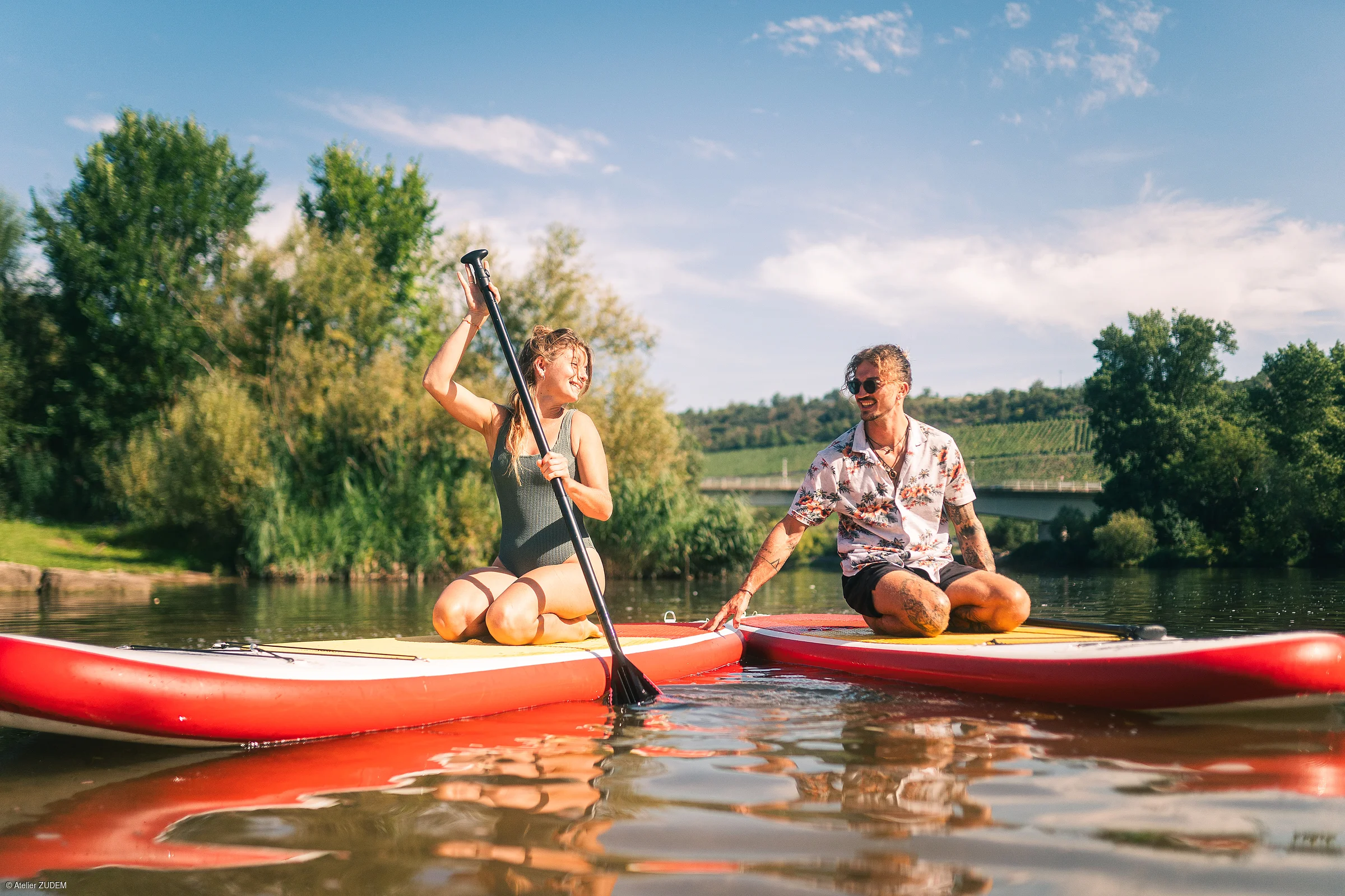 Zwei Personen sitzen auf Stand-up-Paddle-Boards auf einem ruhigen Fluss, umgeben von Bäumen und blauem Himmel.