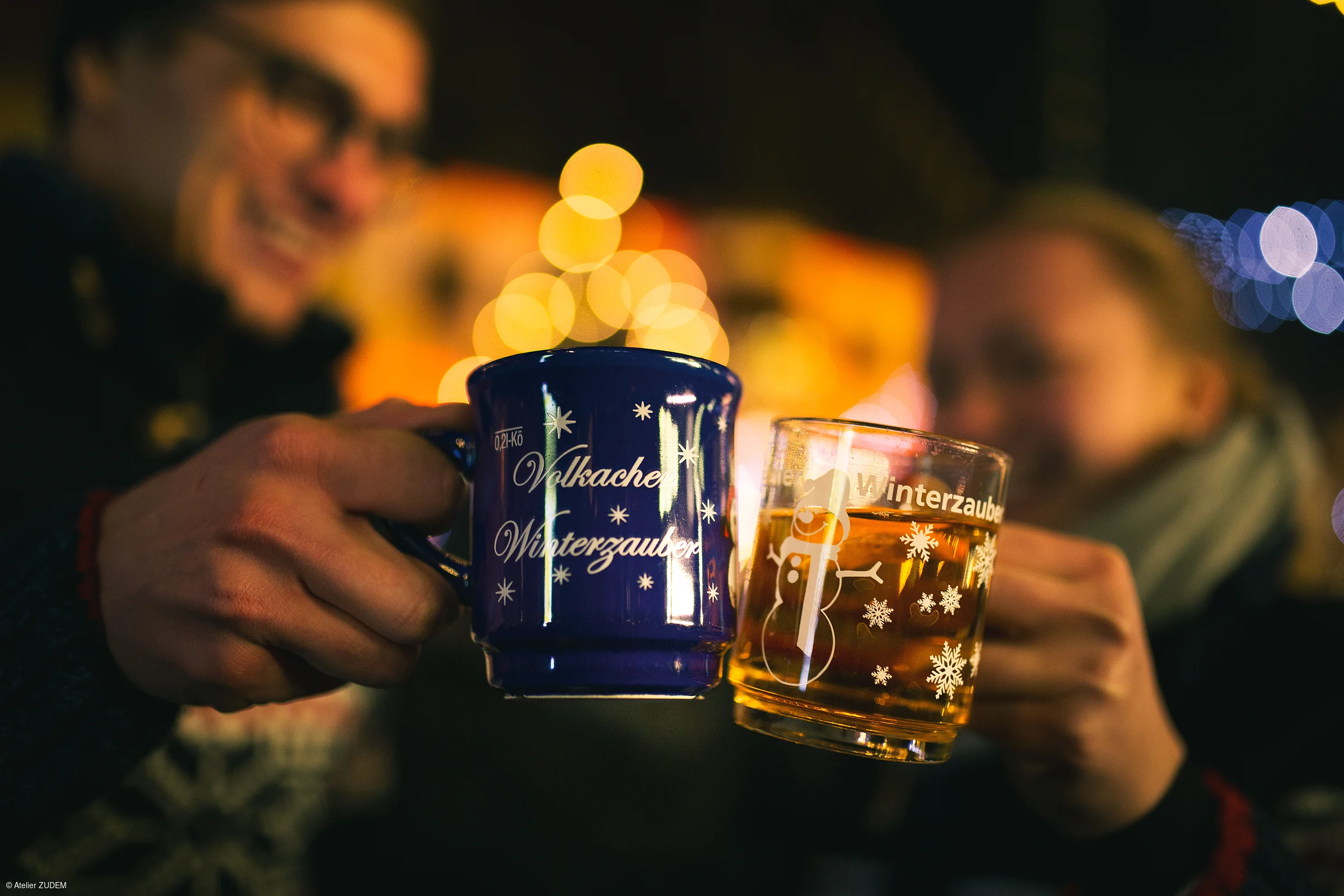 Zwei Personen stoßen mit Tassen an, eine blaue und eine transparente mit Wintermotiven, unscharfer Hintergrund.