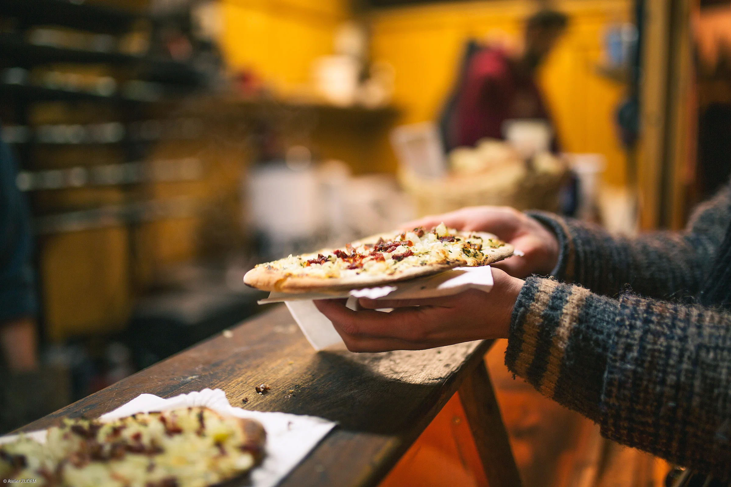 Person hält eine belegte Flammkuchen-Scheibe auf Serviette über einem Holztisch in einer warm beleuchteten Umgebung