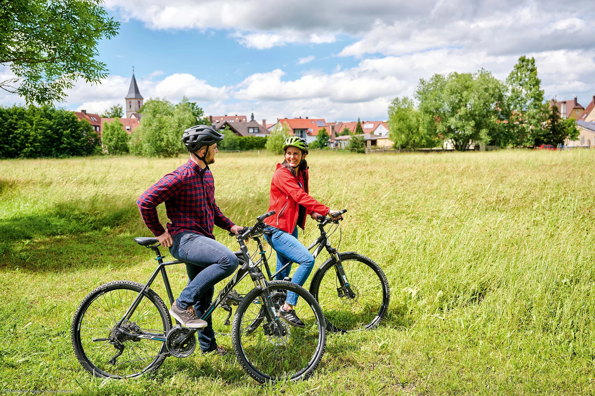 Zwei Personen mit Fahrradhelmen auf Mountainbikes auf einer Wiese vor einem Dorf mit Häusern und Kirche im Hintergrund.