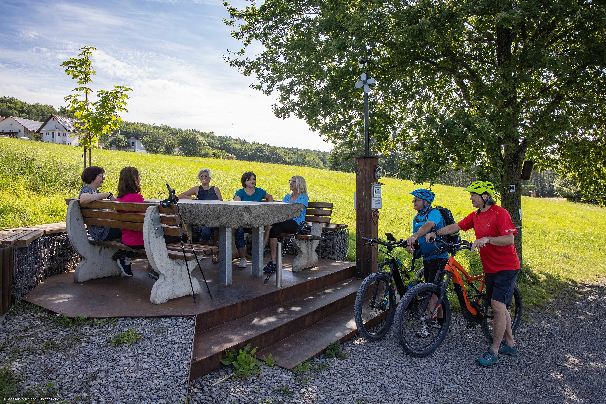 Sechs Frauen sitzen an einem runden Tisch auf einer Bank, zwei Männer mit Fahrrädern stehen daneben unter einem Baum.