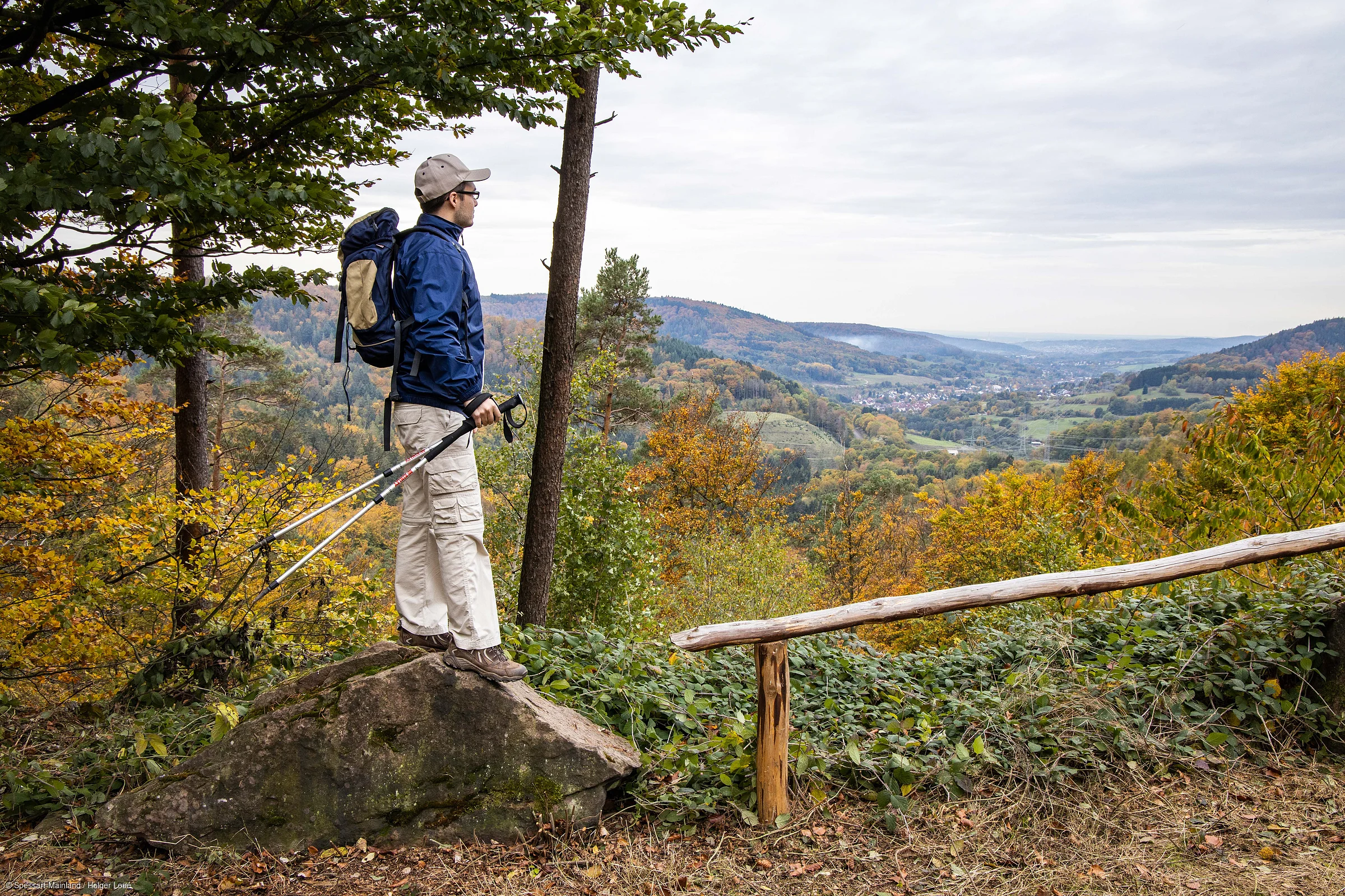 Wanderer mit Rucksack und Stöcken steht auf Felsen und blickt auf herbstliche Landschaft mit Hügeln und Tal.