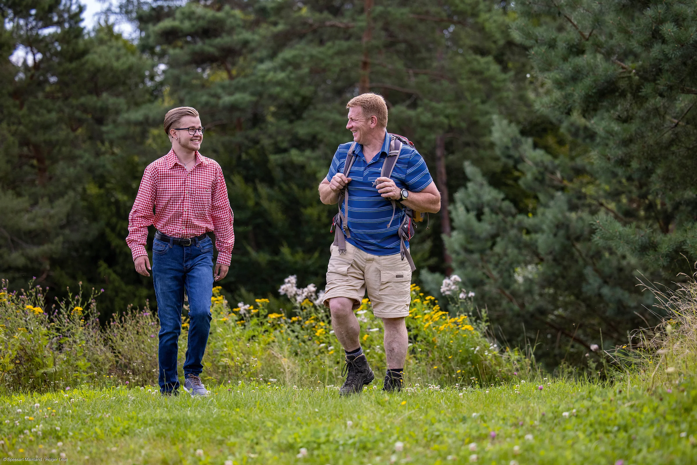 Zwei Männer wandern auf einem Wiesenweg, umgeben von Bäumen und Wildblumen.