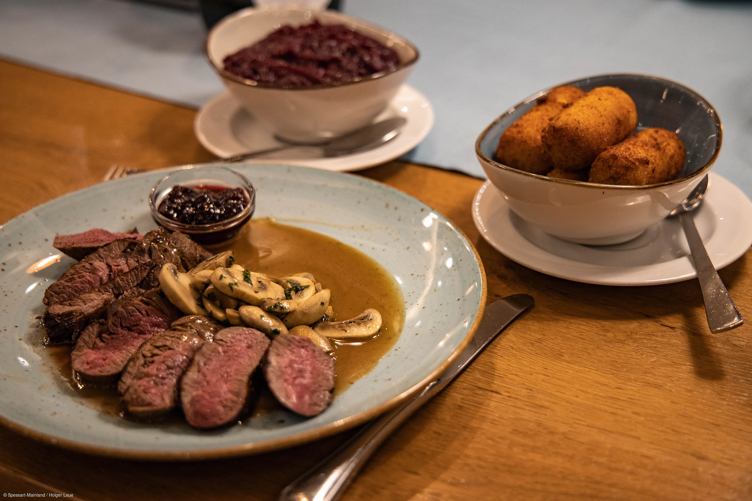 Teller mit gebratenem Fleisch, Pilzen und Sauce, daneben Schale mit Kroketten und Schale mit Rotkohl auf Holztisch