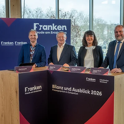 Vier Personen stehen an einem Holztisch mit Beschriftung „Bilanz und Ausblick 2026 Pressegespräch in Ochsenfurt“ vor blauem Hintergrund mit Franken-Logo.