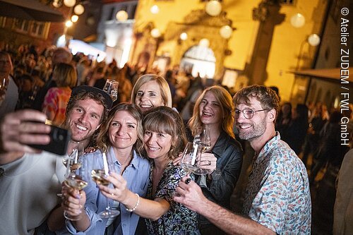 Pressereise Fränkisches Weinland Sechs Personen mit Weingläsern nehmen bei Nacht auf einem Fest ein Selfie auf, im Hintergrund viele Gäste.