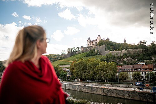 Pressereise Würzburg Frau mit rotem Schal blickt auf die Festung Marienberg über dem Main in Würzburg bei Tageslicht.
