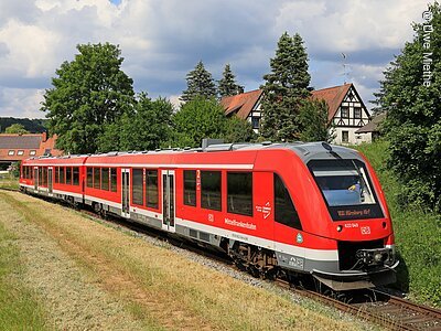 Roter Regionalzug der Mittelfrankenbahn fährt auf Gleisen durch eine ländliche Gegend mit Häusern und Bäumen.
