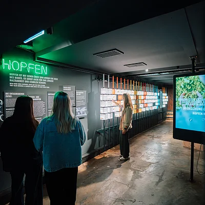 Personen betrachten eine Ausstellung über Hopfen in einem dunklen Raum mit beleuchteten Tafeln und einem Bildschirm.