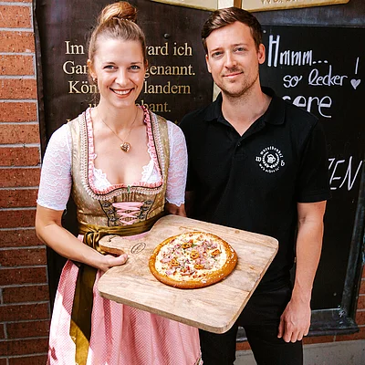 Zwei Personen stehen vor einer Backsteinwand. Eine Frau in Tracht hält ein Holzbrett mit einer Brizza.