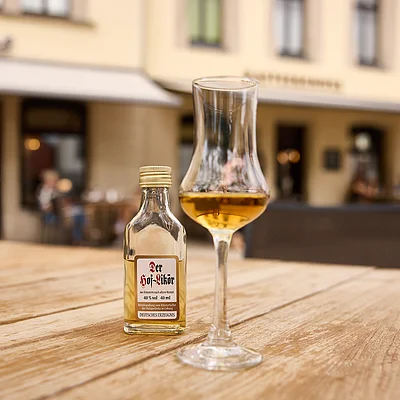Ein Glas und eine kleine Flasche mit Likör stehen auf einem Holztisch im Freien vor einem Gebäude.