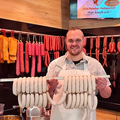 Mann mit Schürze hält zwei Stangen mit Weißürsten vor einer Theke mit hängenden Würsten.