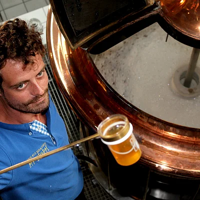 Mann in blauem Hemd hält Probe mit goldenem Bier vor kupferfarbenem Braukessel mit Schaum.