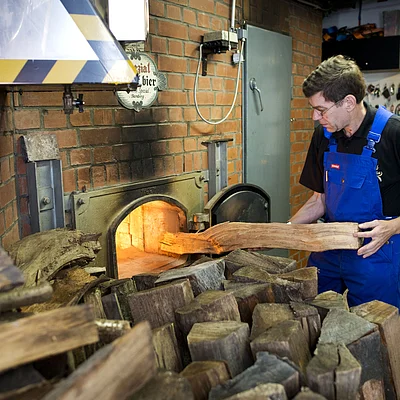 Person in blauer Latzhose legt Holzscheit in einen Ofen in einem Backsteingebäude.