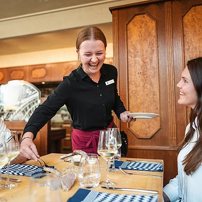 Kellnerin serviert Essen an Tisch mit drei Gästen in Restaurant mit Holzmöbeln und Weingläsern.