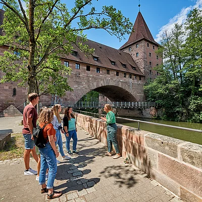 Fünf Personen stehen an einer Steinmauer an einem Fluss vor einer historischen Brücke mit Turm bei sonnigem Wetter.