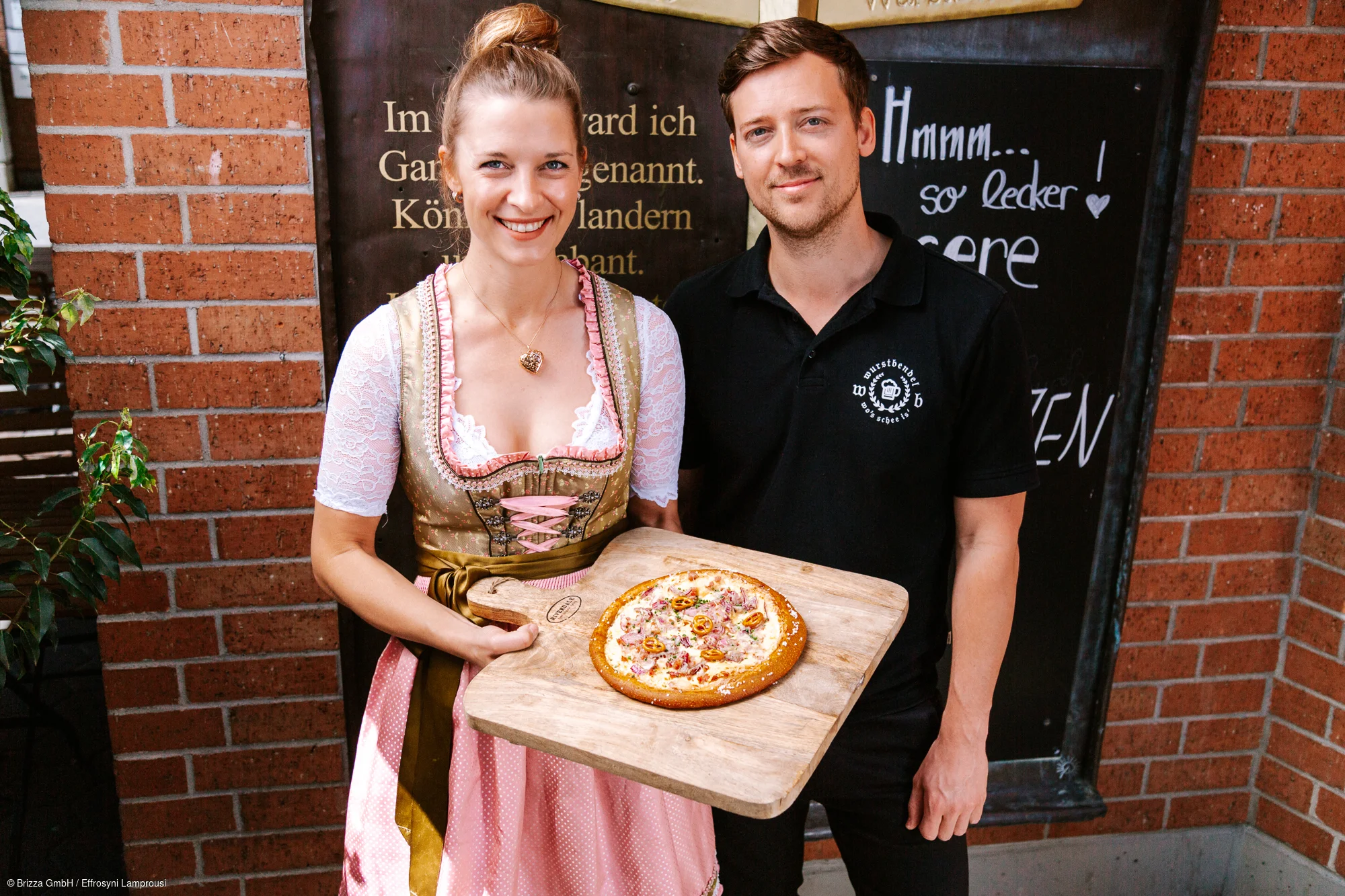Zwei Personen stehen vor einer Backsteinwand. Eine Frau in Tracht hält ein Holzbrett mit einer Brizza.