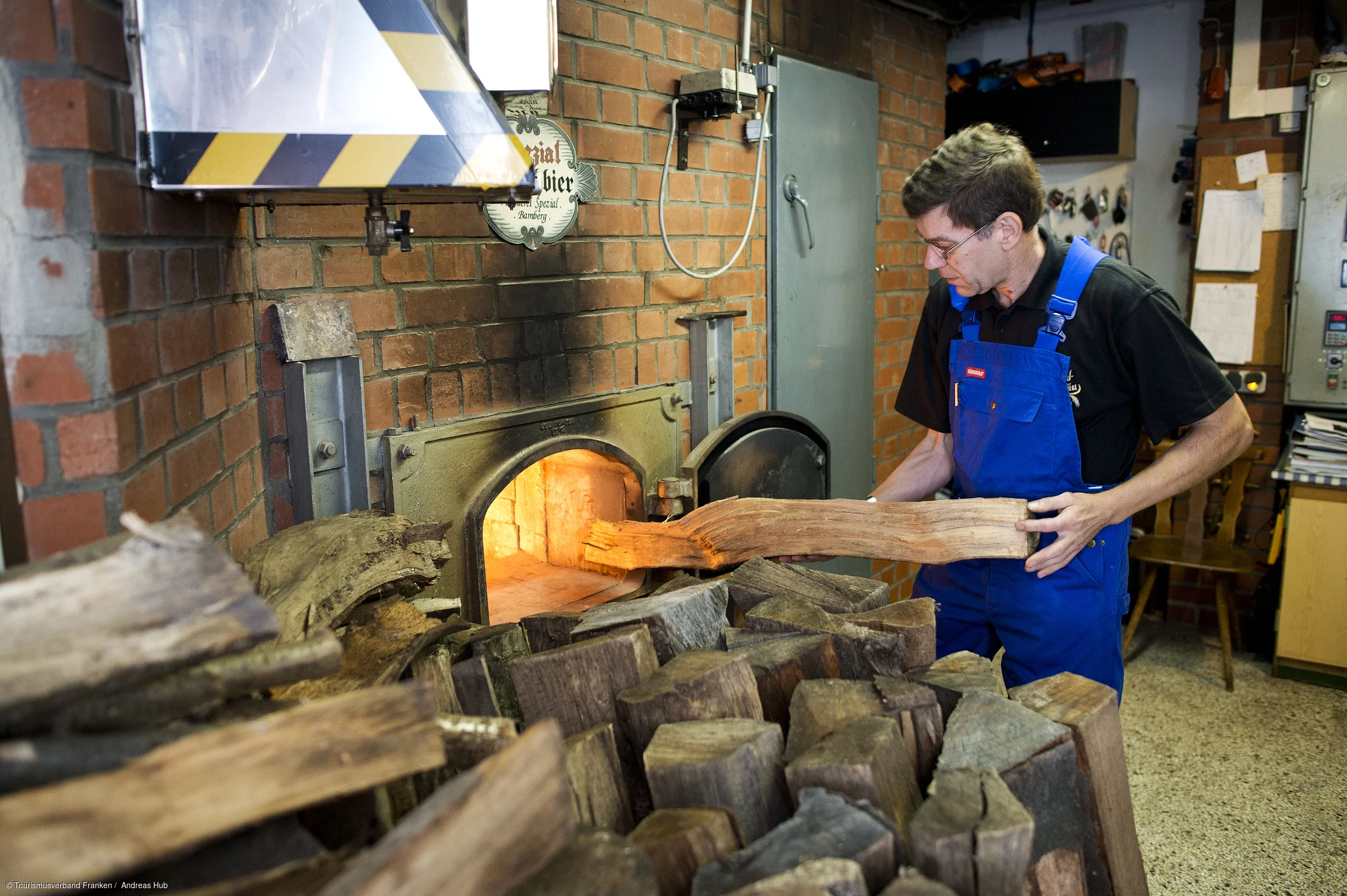 Person in blauer Latzhose legt Holzscheit in einen Ofen in einem Backsteingebäude.