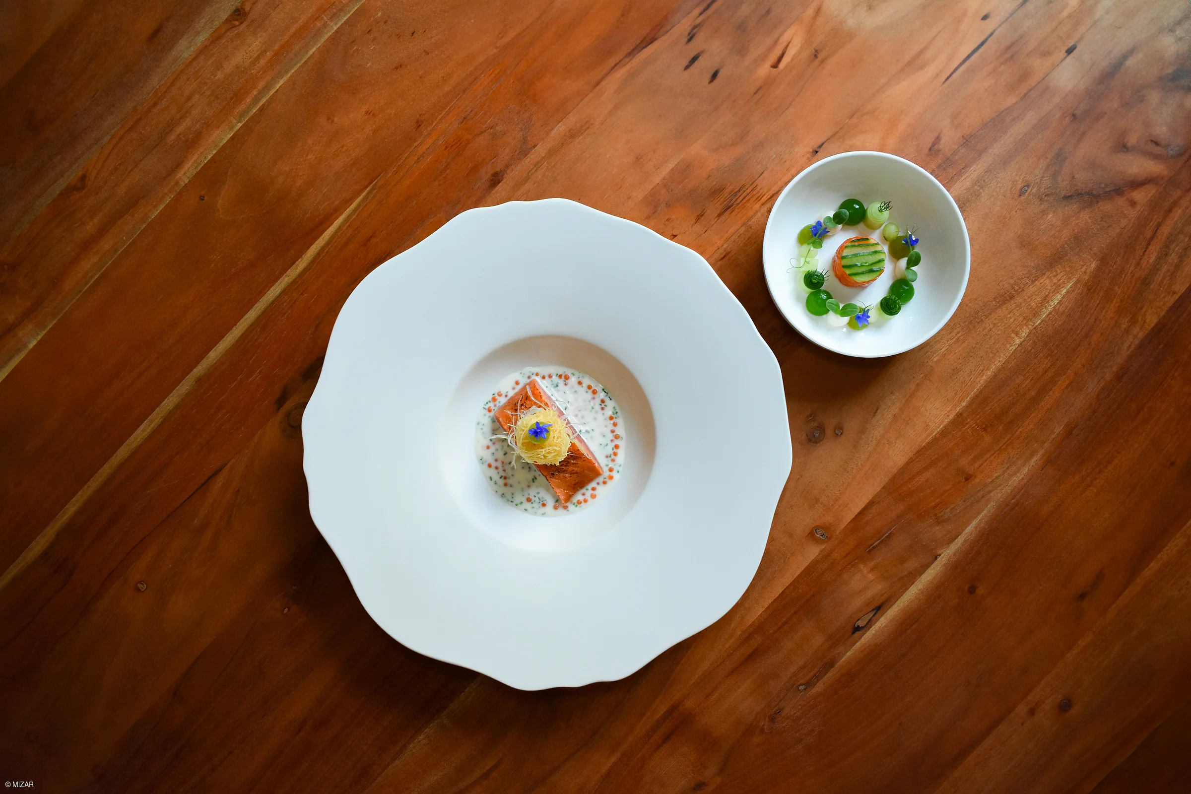 Zwei Teller mit kunstvoll angerichteten Speisen auf einem Holztisch, ein großer und ein kleiner Teller.