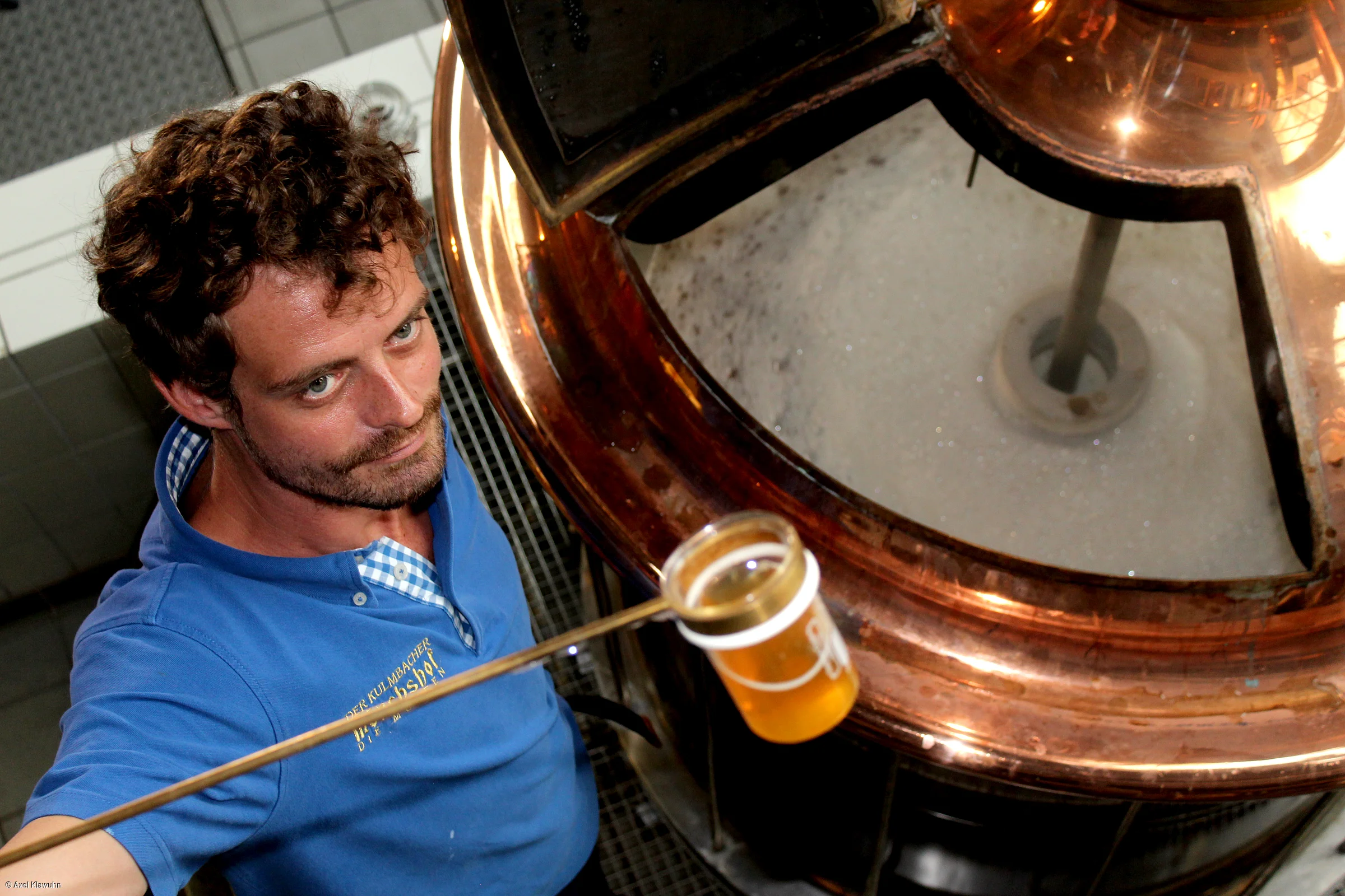 Mann in blauem Hemd hält Probe mit goldenem Bier vor kupferfarbenem Braukessel mit Schaum.