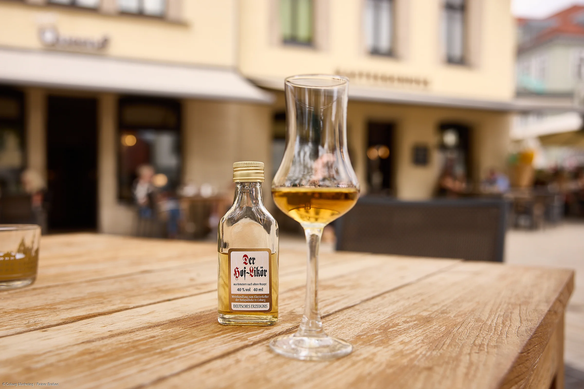 Ein Glas und eine kleine Flasche mit Likör stehen auf einem Holztisch im Freien vor einem Gebäude.