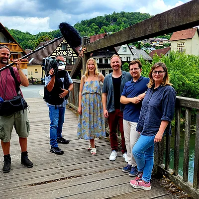 Sechs Personen, darunter ein Kameramann und ein Tonmann, stehen auf einer Holzbrücke vor Fachwerkhäusern und grünen Hügeln.
