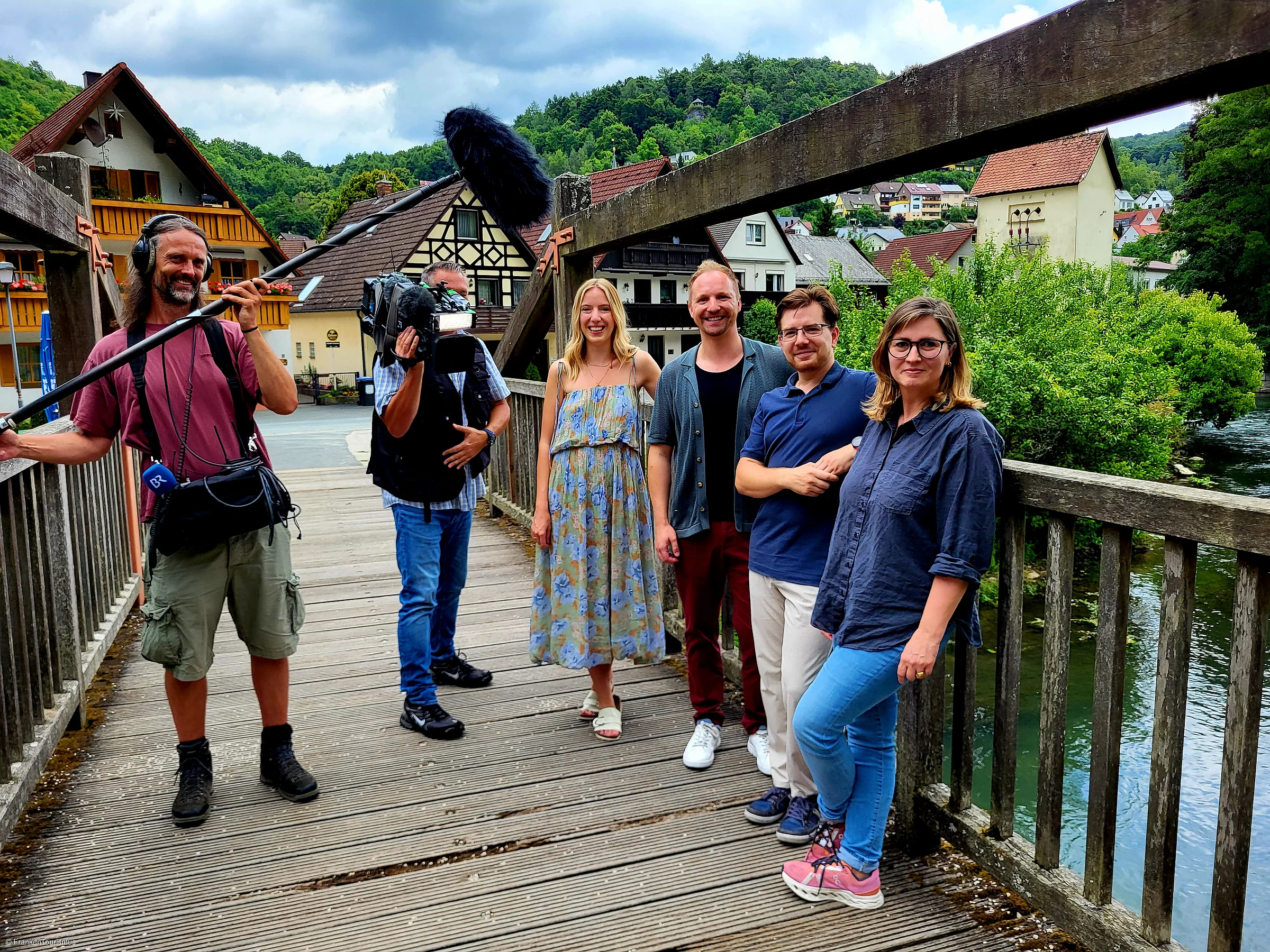 Sechs Personen, darunter ein Kameramann und ein Tonmann, stehen auf einer Holzbrücke vor Fachwerkhäusern und grünen Hügeln.