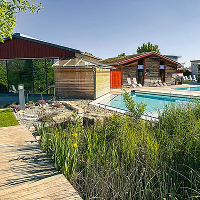 Außenbereich mit Schwimmbecken, Liegestühlen, Holzsteg und Gebäuden unter klarem Himmel.